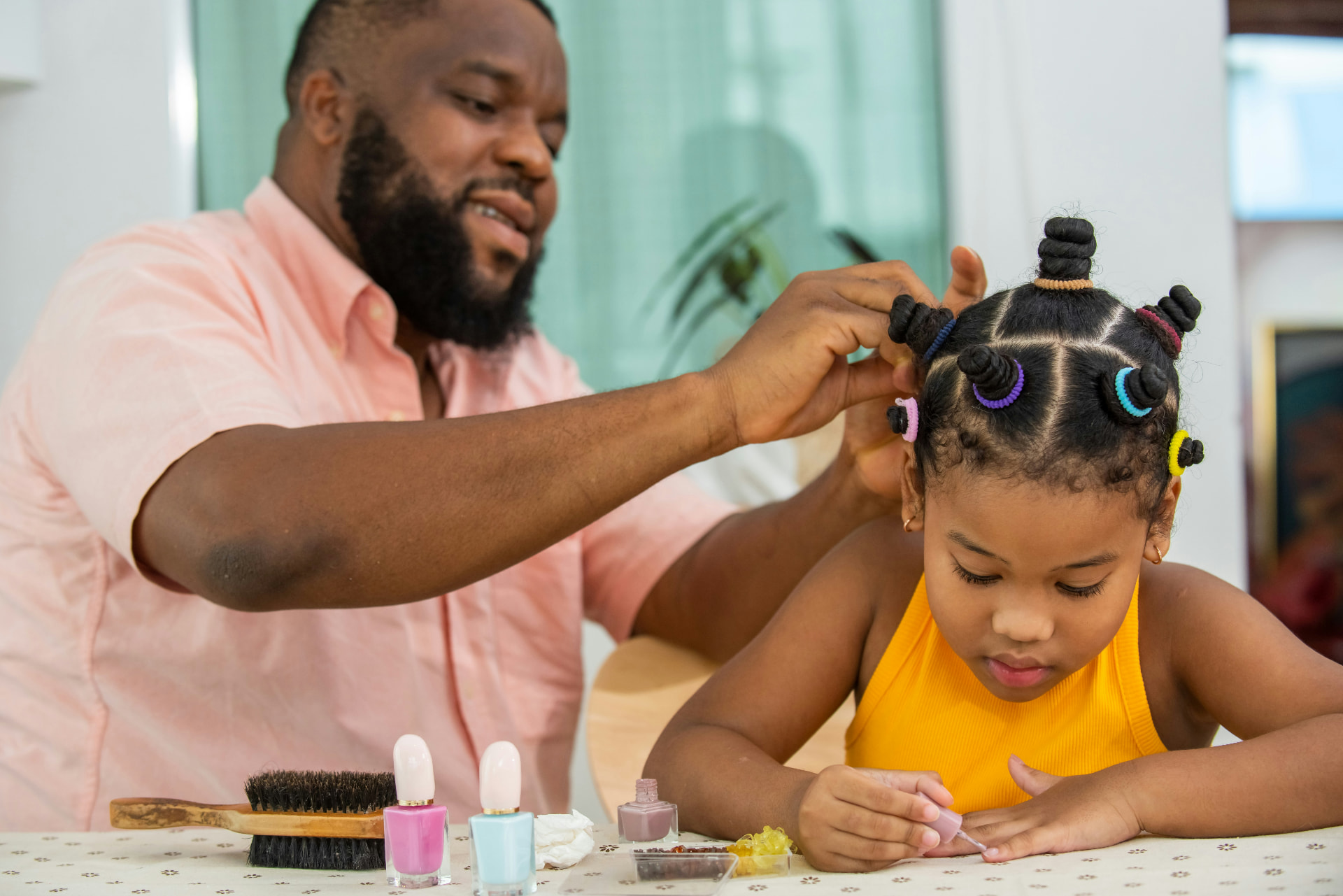 An adult styling a child's hair at a table while the child paints her nails